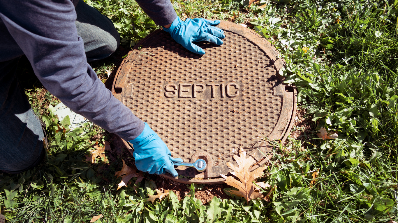 Person in blue gloves opens a septic tank cover in a grassy area.