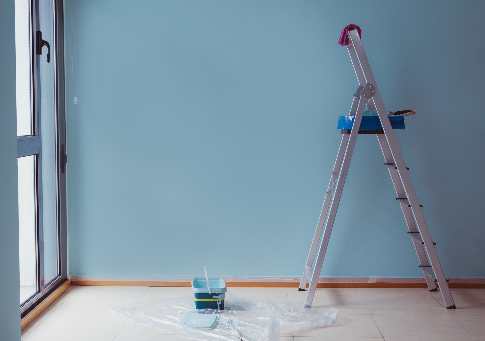 Room with a light blue wall, ladder, and paint supplies on the floor.