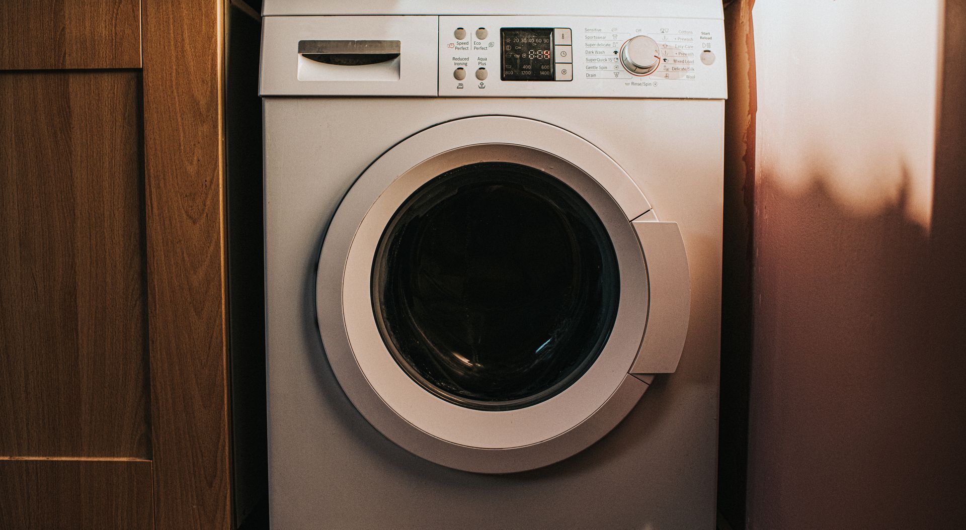 White washing machine in a small space, between a wooden door and a pink wall.