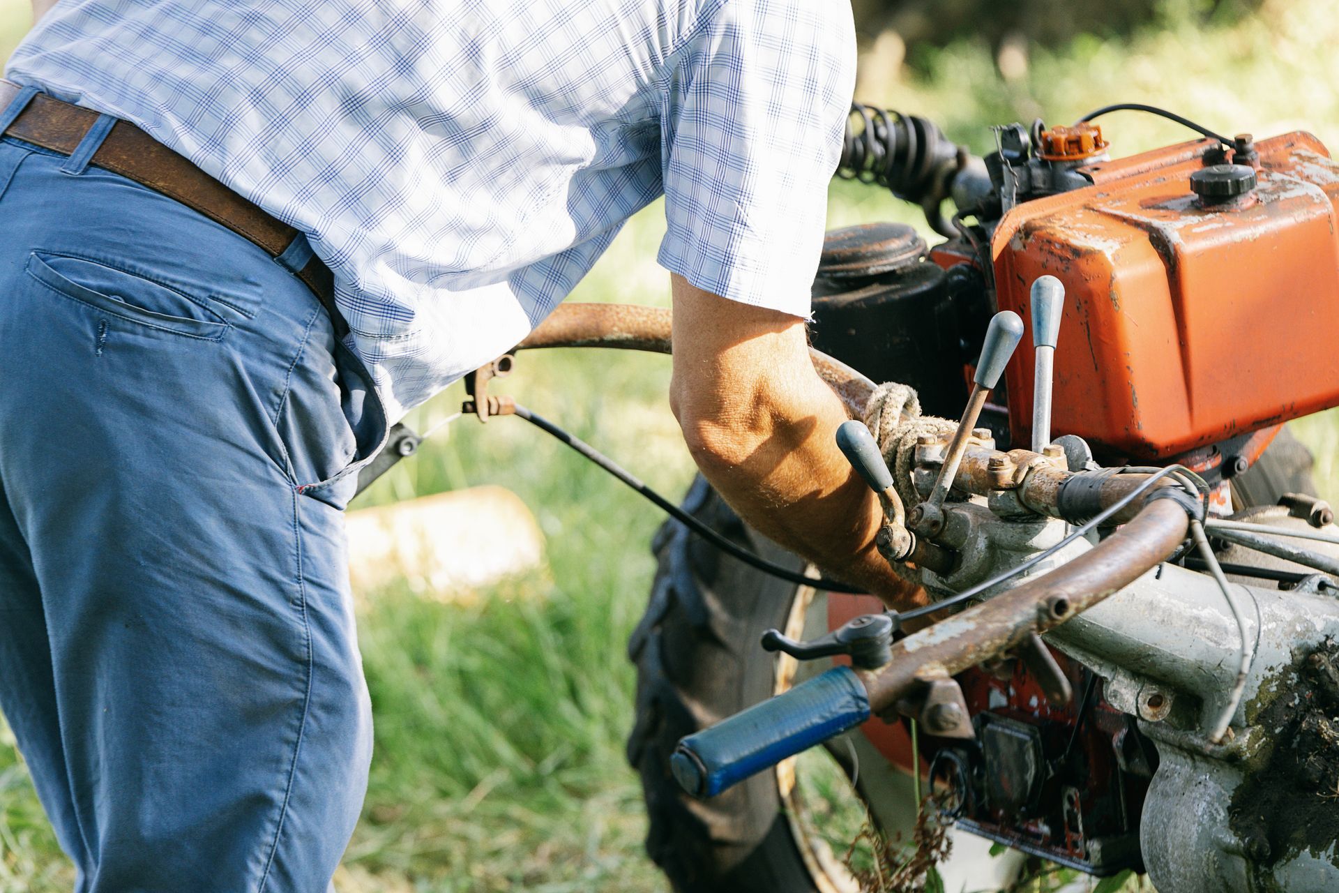 A man in blue jeans and a white shirt operates a small, red tractor outdoors.