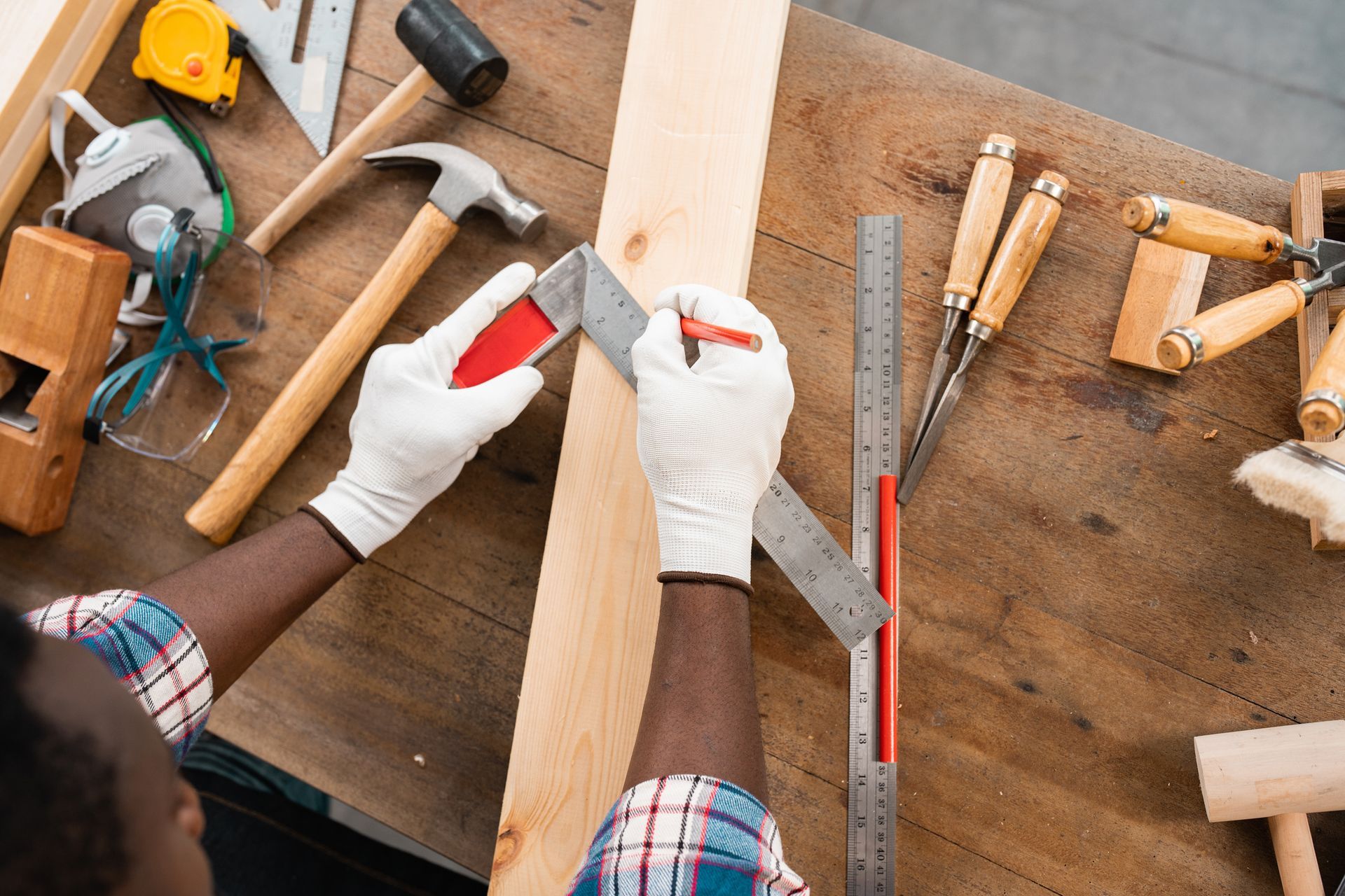 Person wearing gloves using a square to mark a wooden plank on a workbench surrounded by tools.