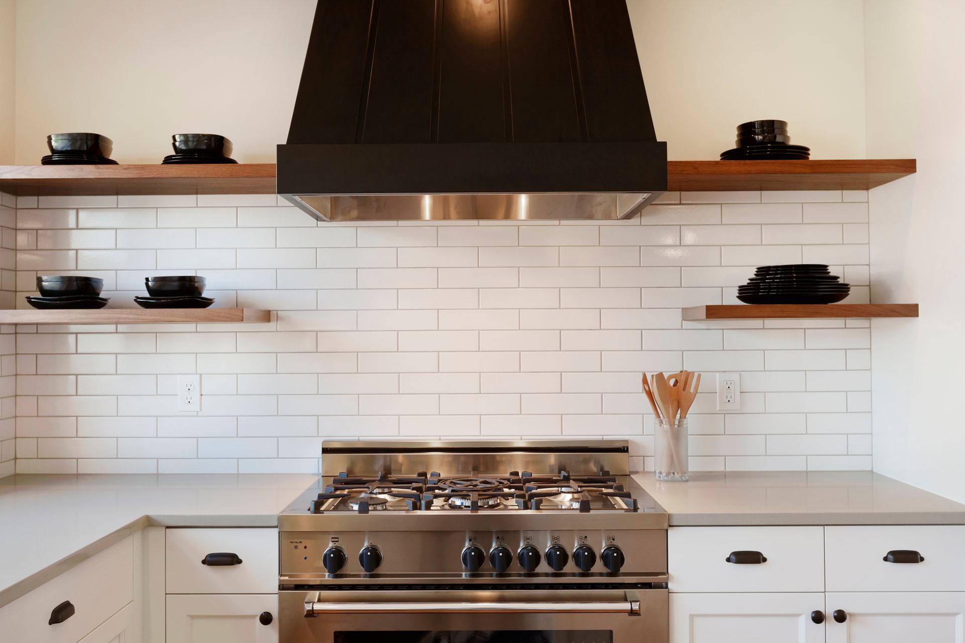 Kitchen with white subway tile backsplash, stainless steel range, black range hood, and floating wooden shelves with black dishes.