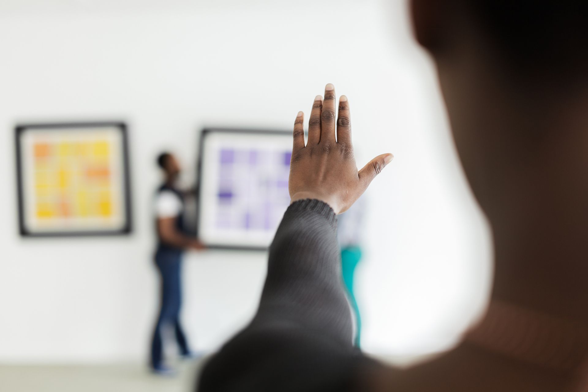 Person's hand raised in front of a blurred art gallery scene; another person hanging artwork.