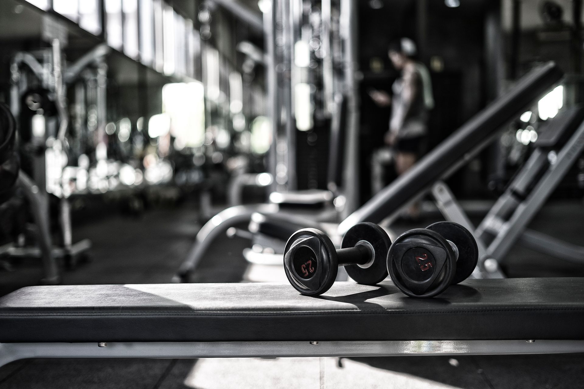 Two dumbbells on a bench in a gym, with workout equipment and a person in the background.