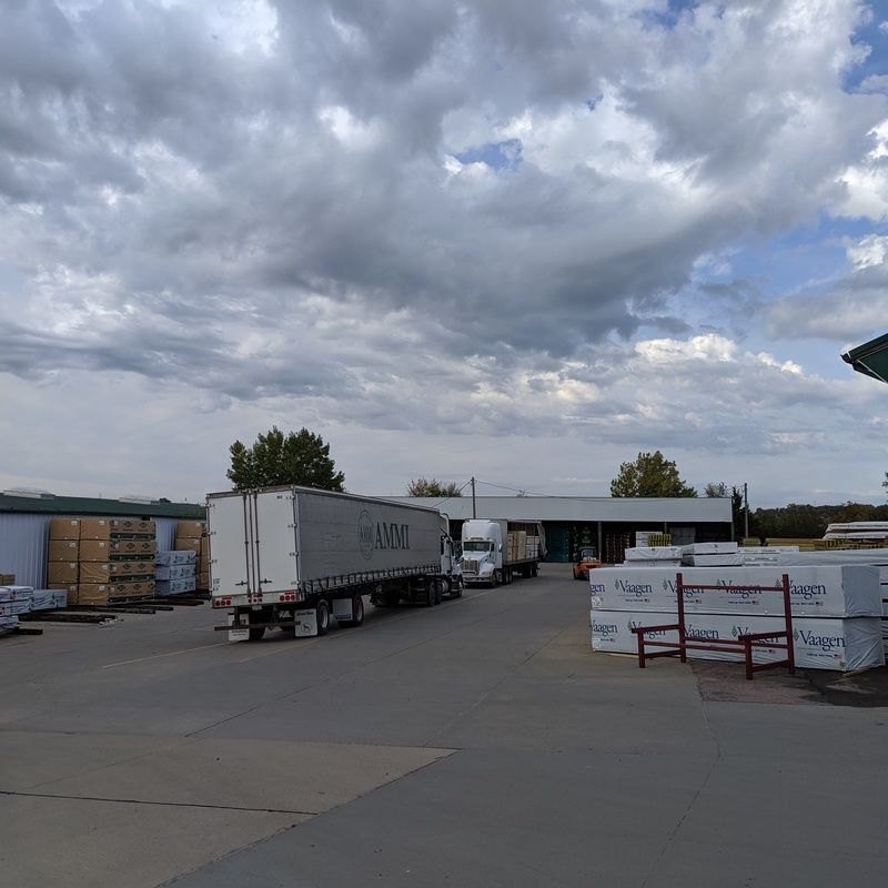 Semi-trucks at a lumberyard under a cloudy sky, loading and unloading wood products.