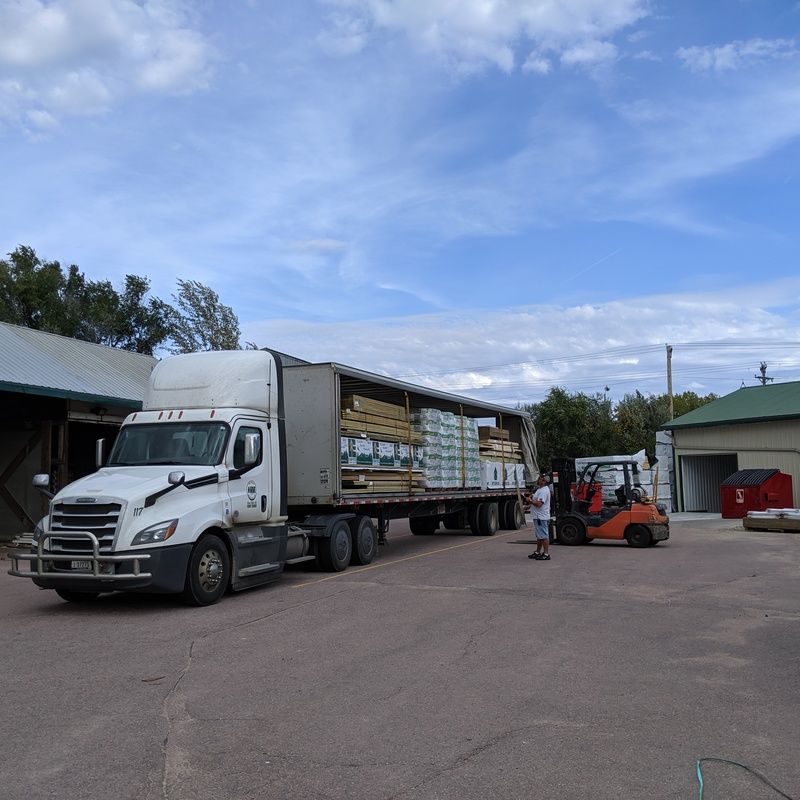 Truck unloading lumber at a building supply store; a forklift moves pallets; blue sky overhead.
