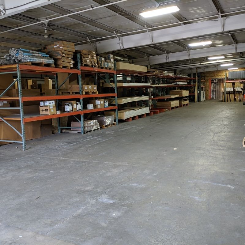 Warehouse interior with metal shelving filled with boxes, lumber, and supplies. Concrete floor, overhead lighting.
