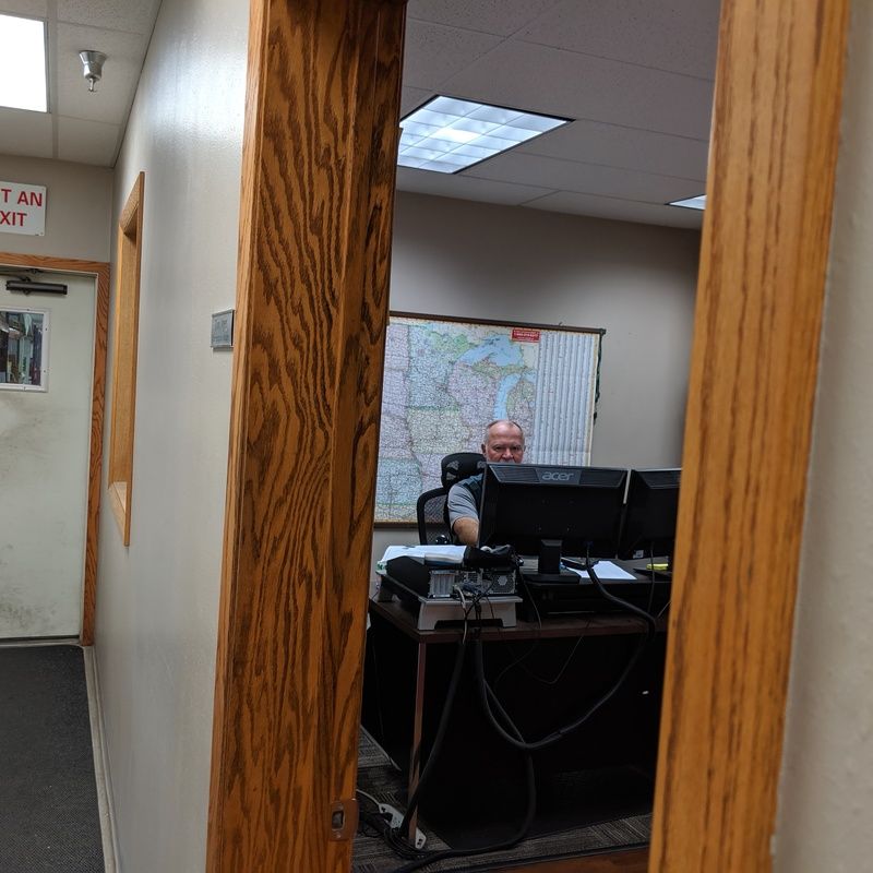 Man at a desk in an office, framed by a doorway. Map on wall, computer monitors, paperwork.
