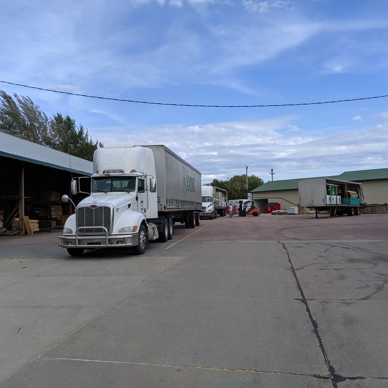 White semi-truck parked in a loading area with several other trucks and buildings under a partly cloudy sky.