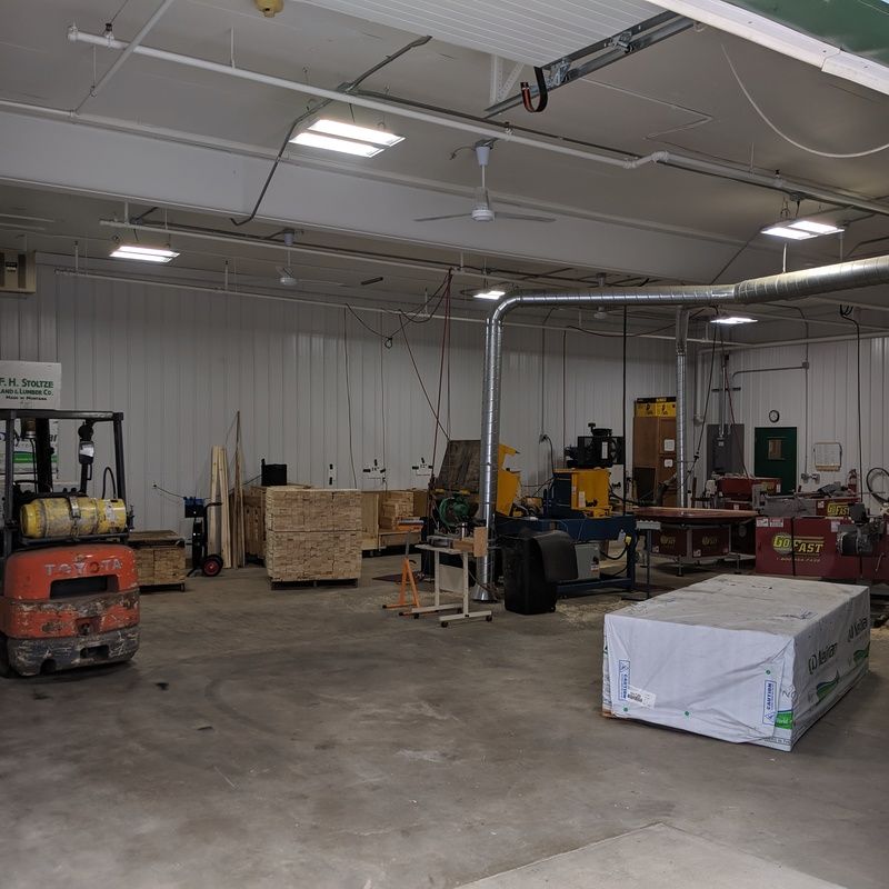 Interior of a warehouse with a forklift, wood pallets, and machinery. Gray concrete floor, white walls.