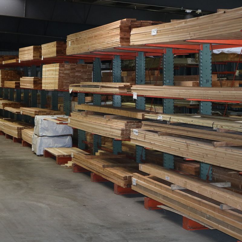 Lumber stacked on shelves in a warehouse. Various sizes and types of wood, with gray shelving and a concrete floor.