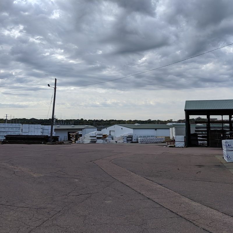 Gray cloudy sky over a lumberyard with white buildings, a dark paved area, and lumber.