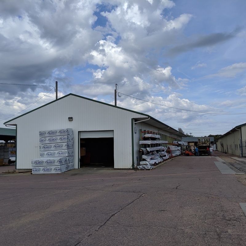 White warehouse with open door and stacks of lumber, under a cloudy sky.