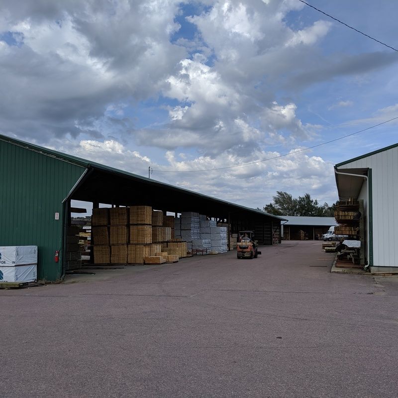 A lumberyard with stacks of wood under a green shed; a forklift operates on the pavement.