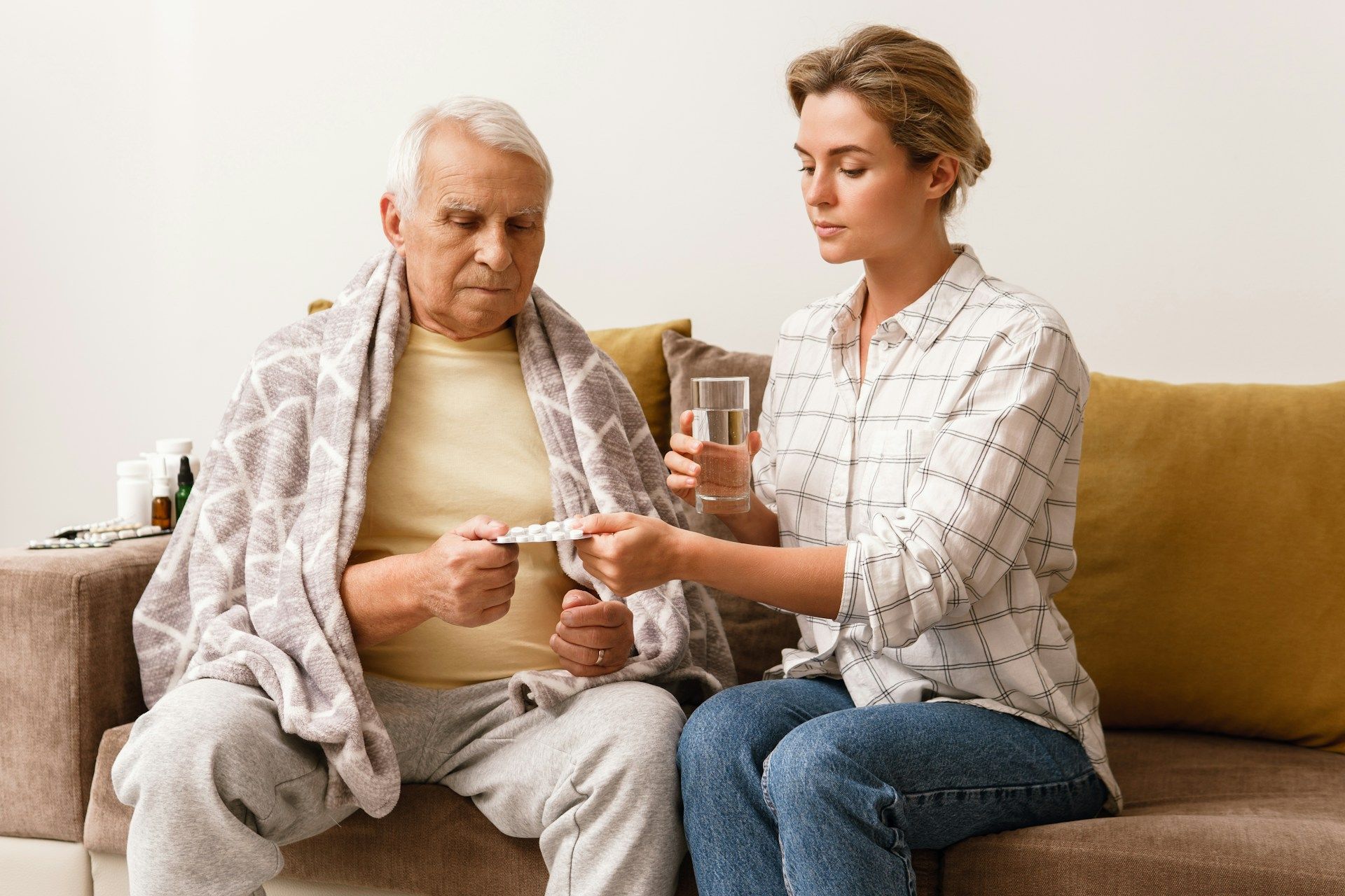Woman giving medication to an older person wrapped in a blanket, sitting on a couch.