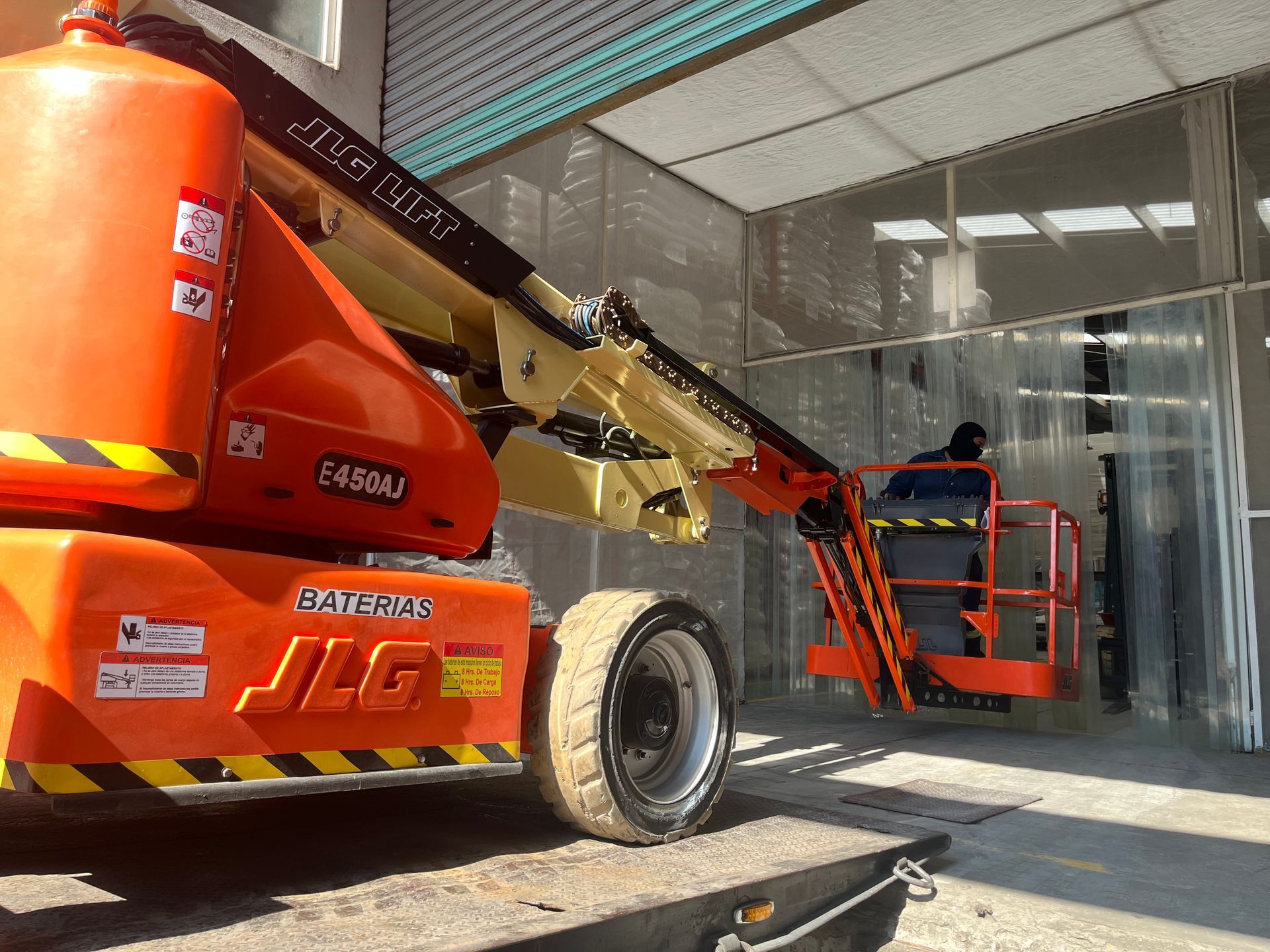 Persona en una plataforma elevadora naranja trabajando cerca de un edificio blanco con detalles rojos.