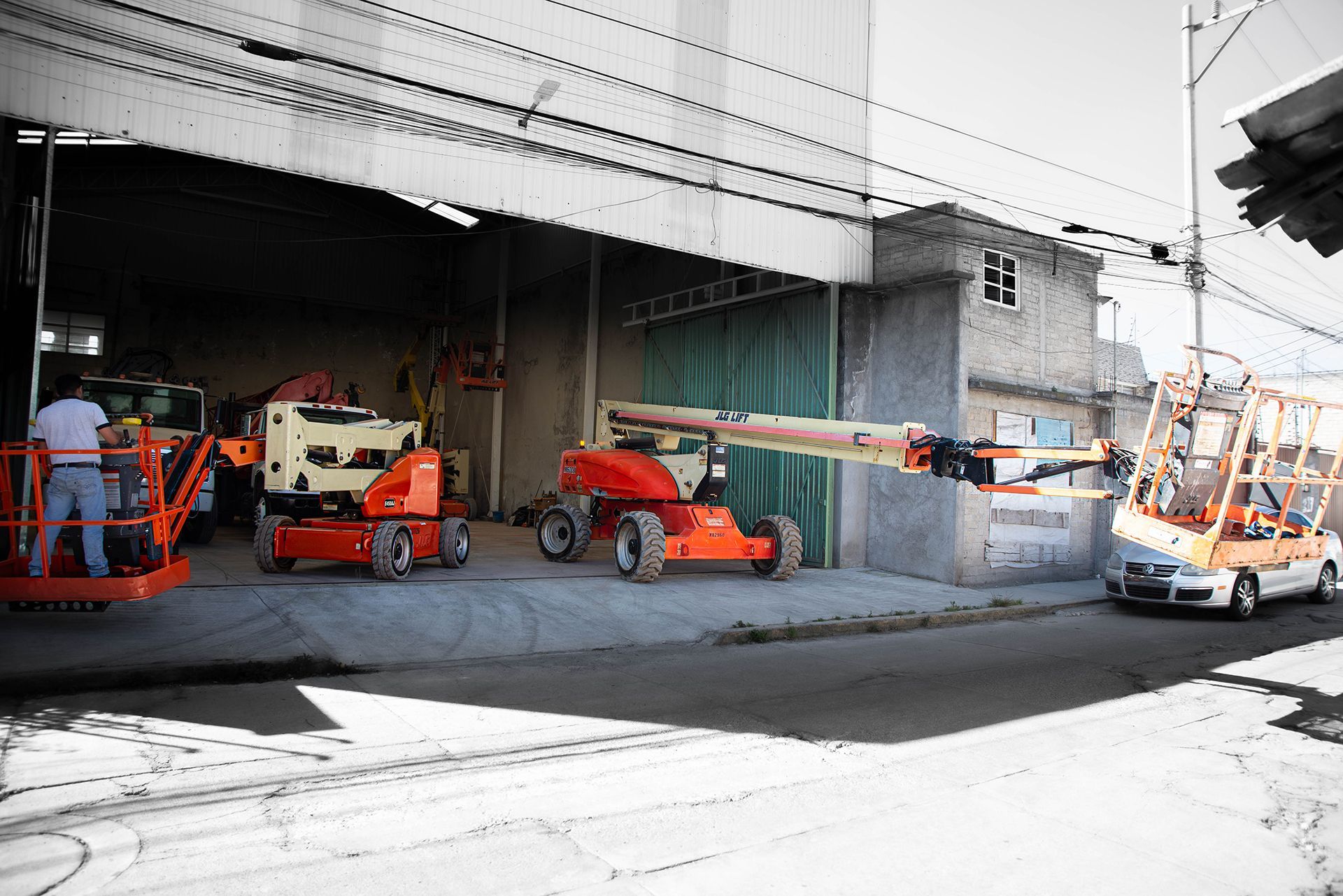 Exterior del almacén; varias plataformas elevadoras de brazo articulado naranjas, una de ellas con el brazo extendido sosteniendo un panel de vidrio junto a una furgoneta de reparto.