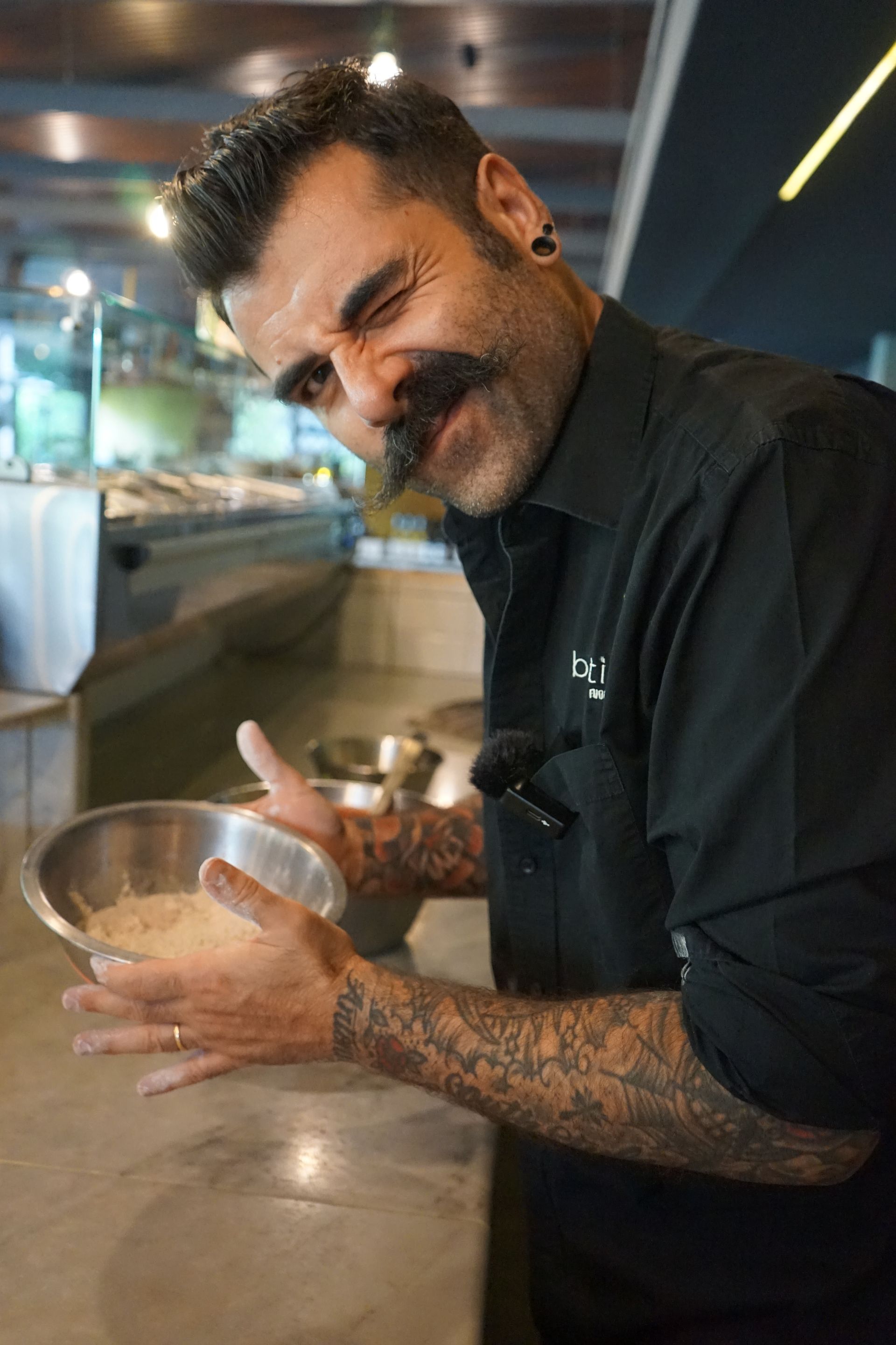 Un uomo con tatuaggi e grandi baffi fa l'occhiolino, tenendo in mano una ciotola, nella cucina di un ristorante.