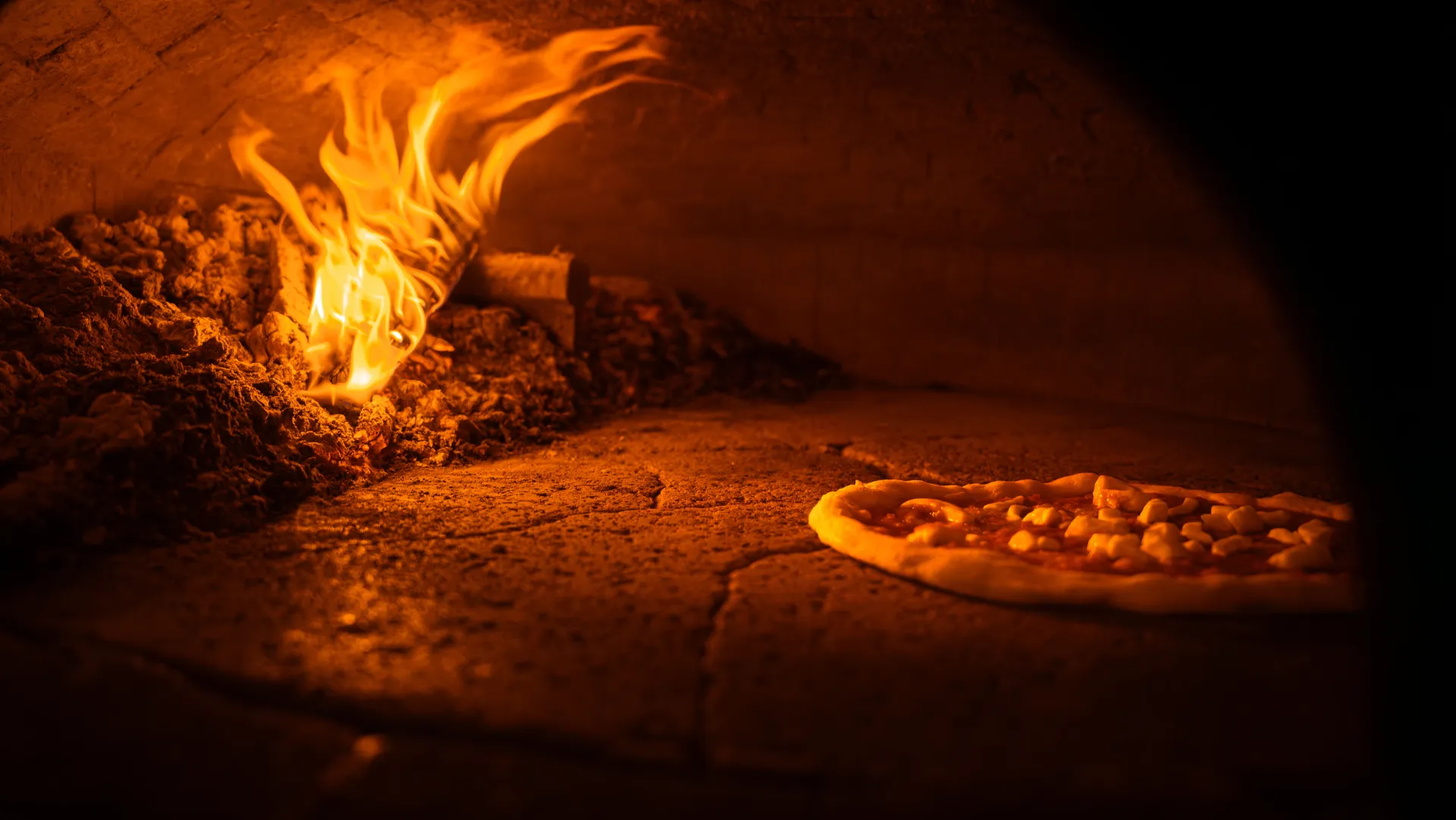 Pizza cotta nel forno a legna con fiamme sullo sfondo.