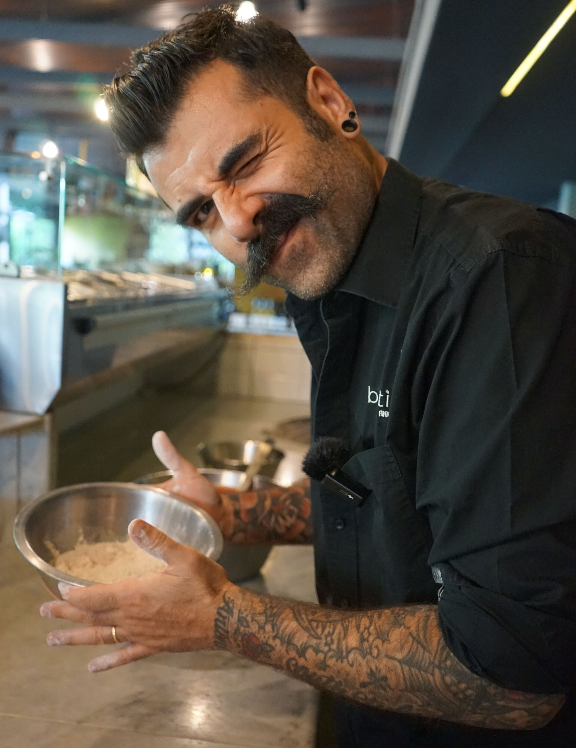 Uomo con baffi e tatuaggi che tiene in mano una ciotola e ammicca, in cucina.