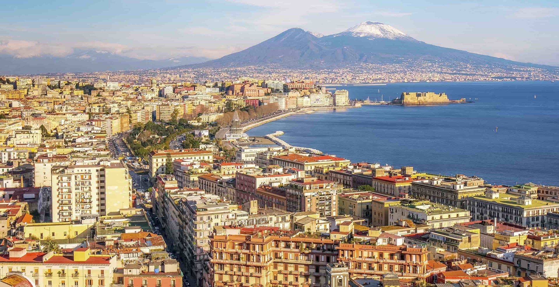 Paesaggio urbano costiero di Napoli, Italia, con il Vesuvio sullo sfondo e un mare azzurro e calmo.