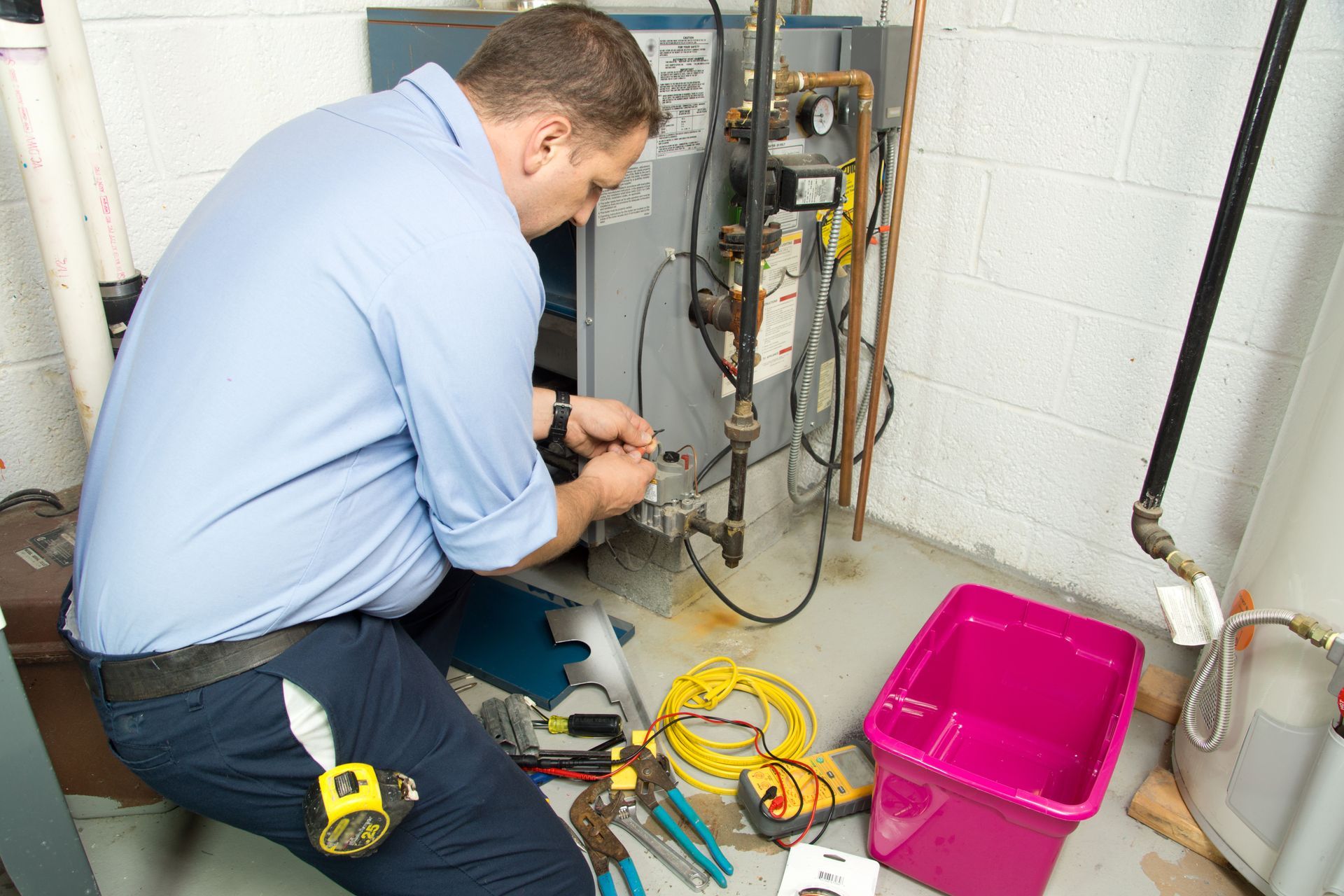Man in blue shirt works on furnace in a basement. Tools and pink bin on the floor.