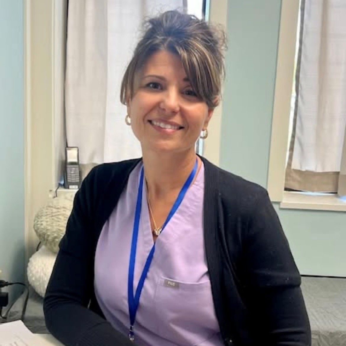 Smiling woman in lavender scrubs and black cardigan with a blue lanyard in a clinic setting
