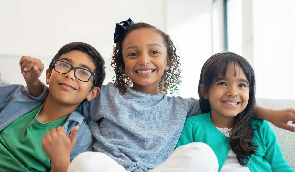 Three children smiling on a couch, leaning together in a bright living room