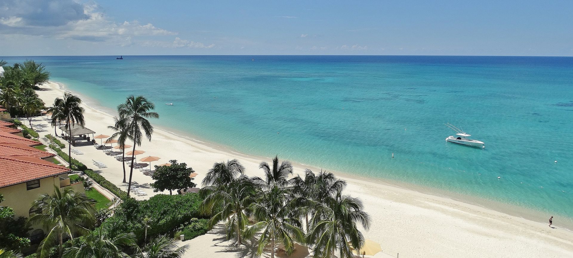Beach in Cayman Islands near one of our properties. Beach with white sand, turquoise water, and palm trees. A small boat is in the distance.