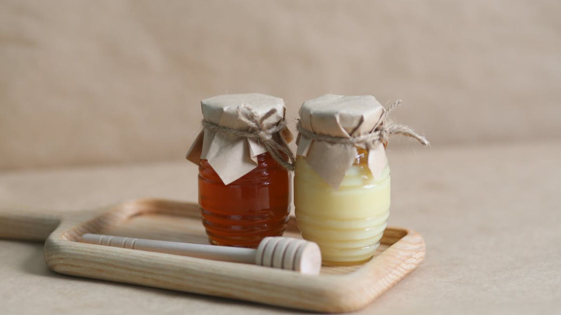 Dos frascos de miel, uno de color ámbar y otro de color crema, con un cucharón de madera para miel sobre una bandeja.