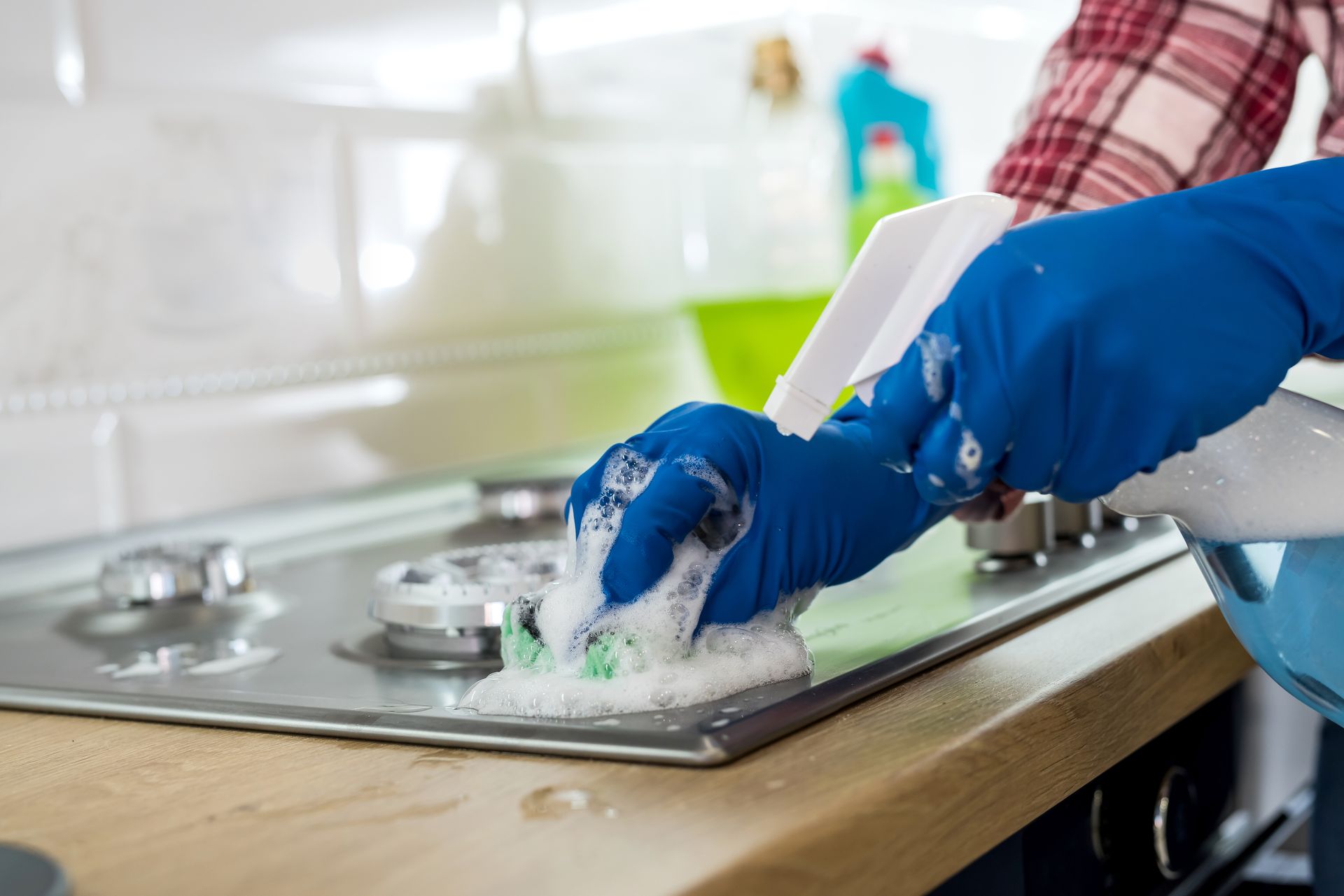 Person wearing blue gloves scrubs a stovetop with a sponge and spray bottle.