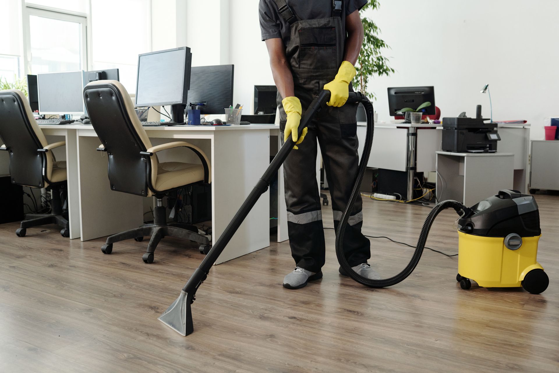 Person in work clothes vacuuming an office floor with a yellow and black vacuum cleaner.