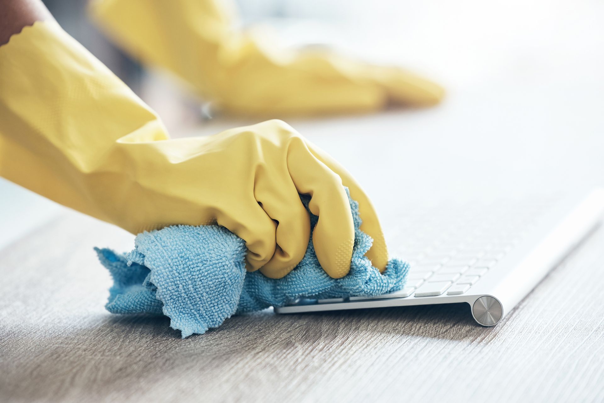 Hands in yellow gloves wiping a keyboard with a blue cloth on a light-colored surface.