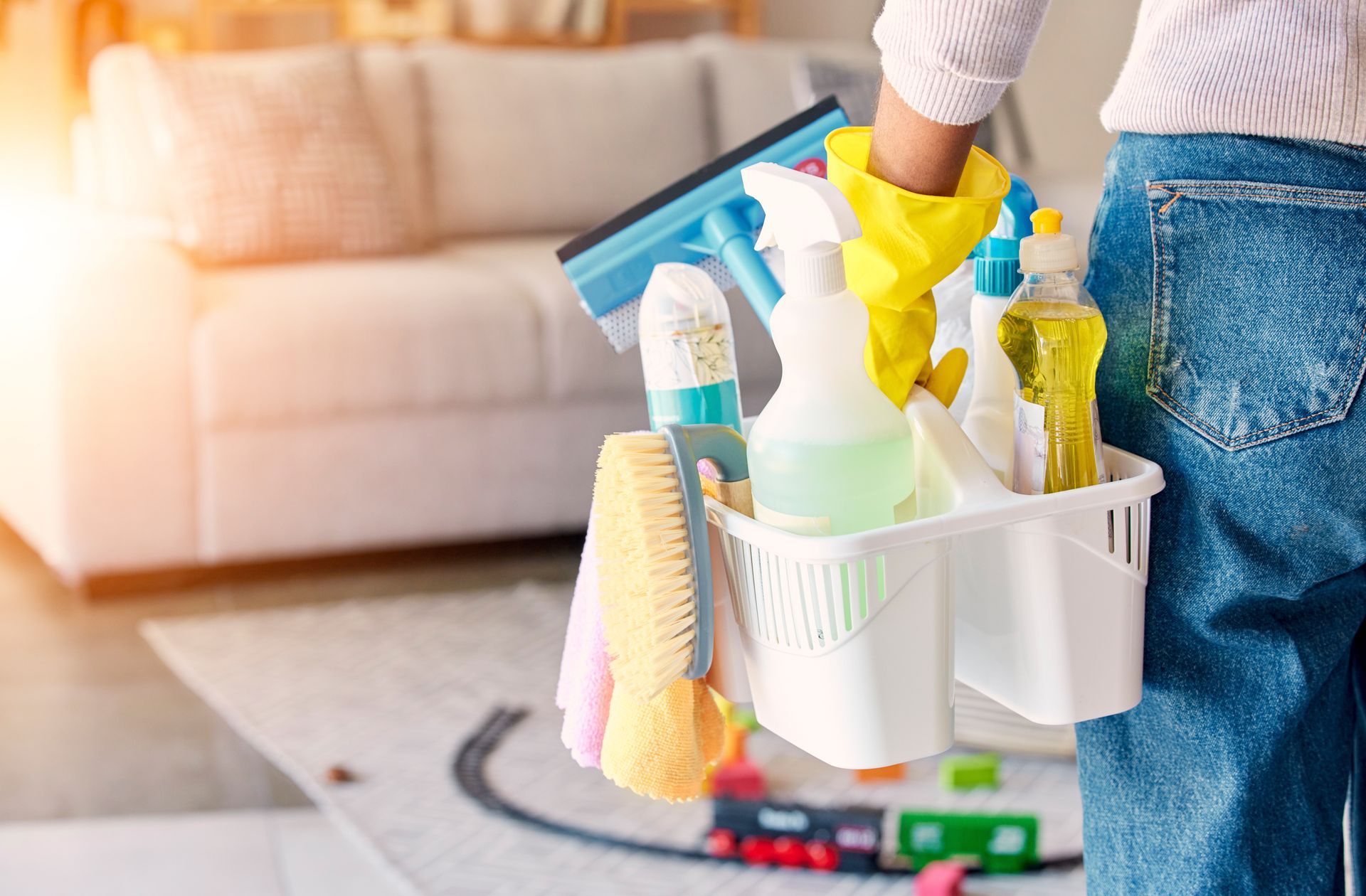 Person holding cleaning supplies in a caddy; yellow gloves, spray bottles, and a brush.