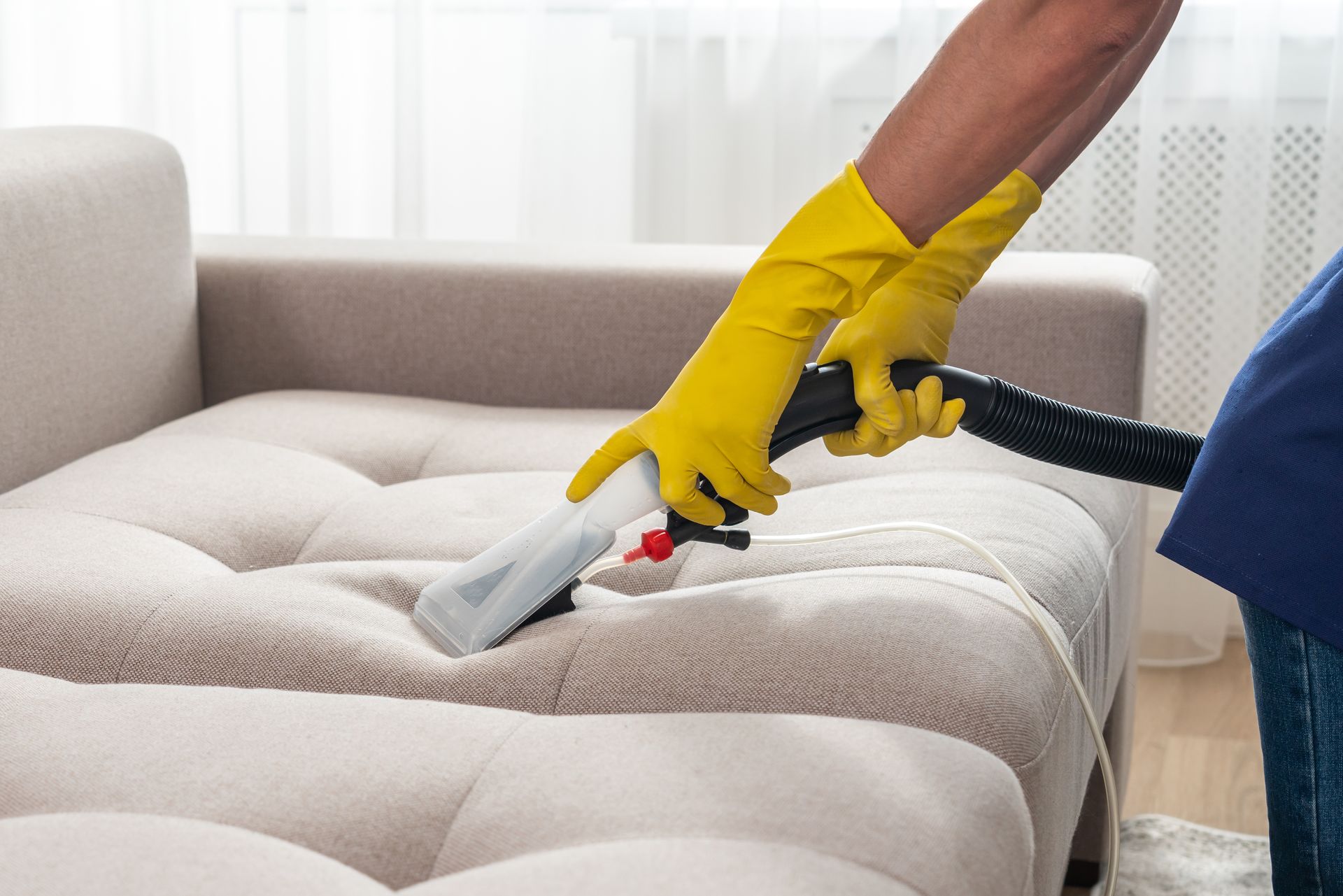 Person wearing yellow gloves cleans a beige sofa with a steam cleaner.