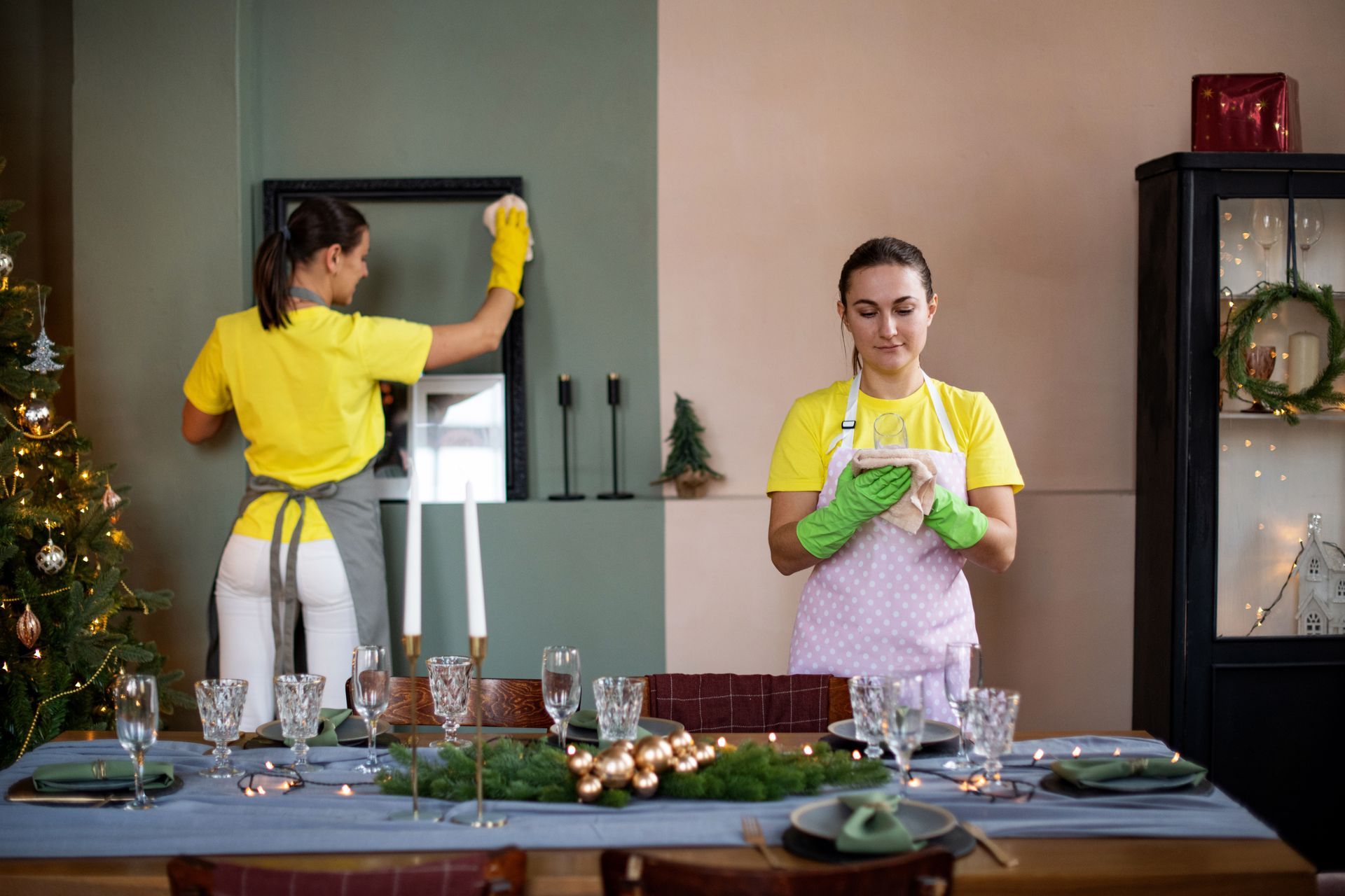 Two women wearing yellow shirts and aprons cleaning a dining room with a Christmas tree.