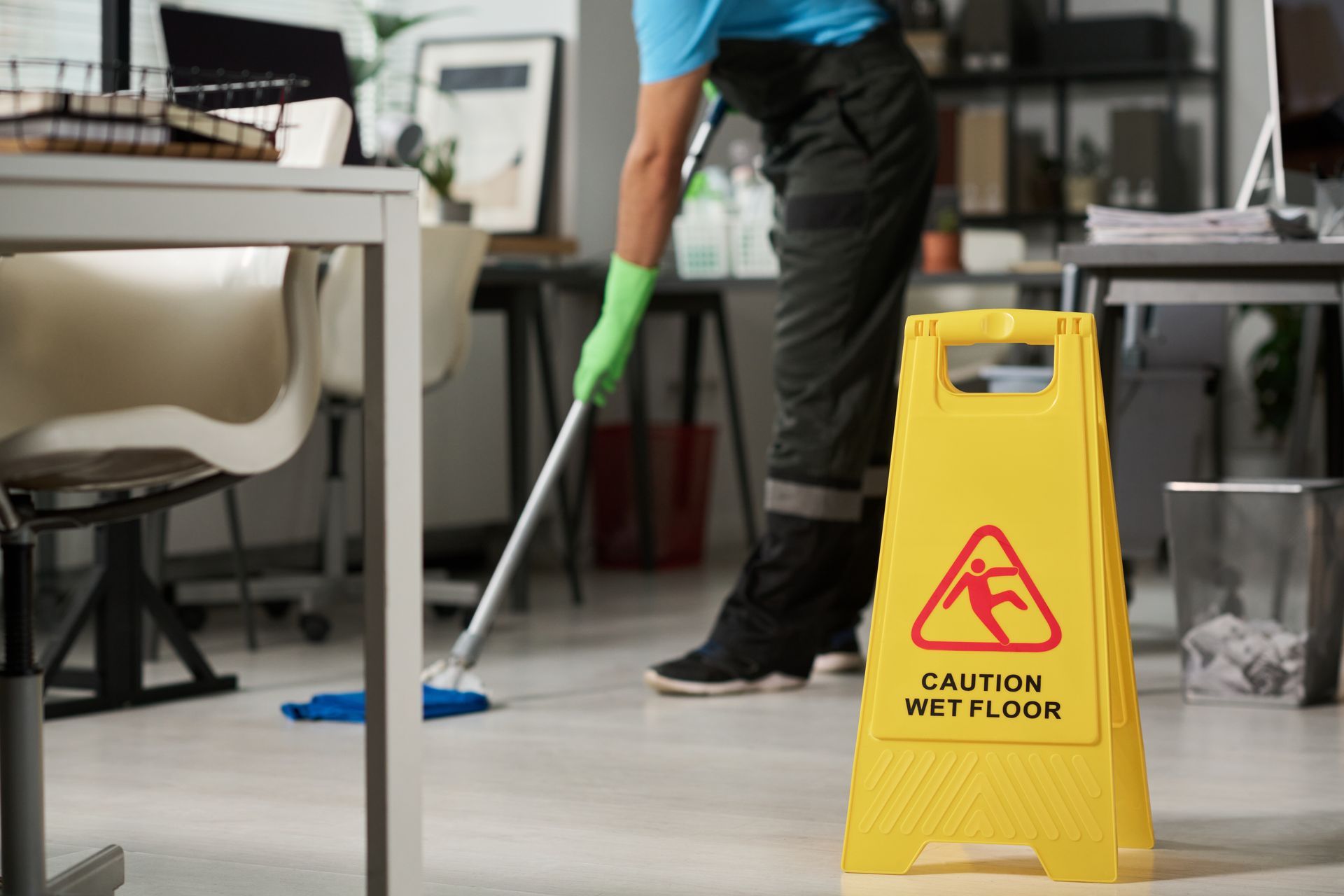 Custodian mopping office floor, yellow caution sign in foreground warns of wet floor.