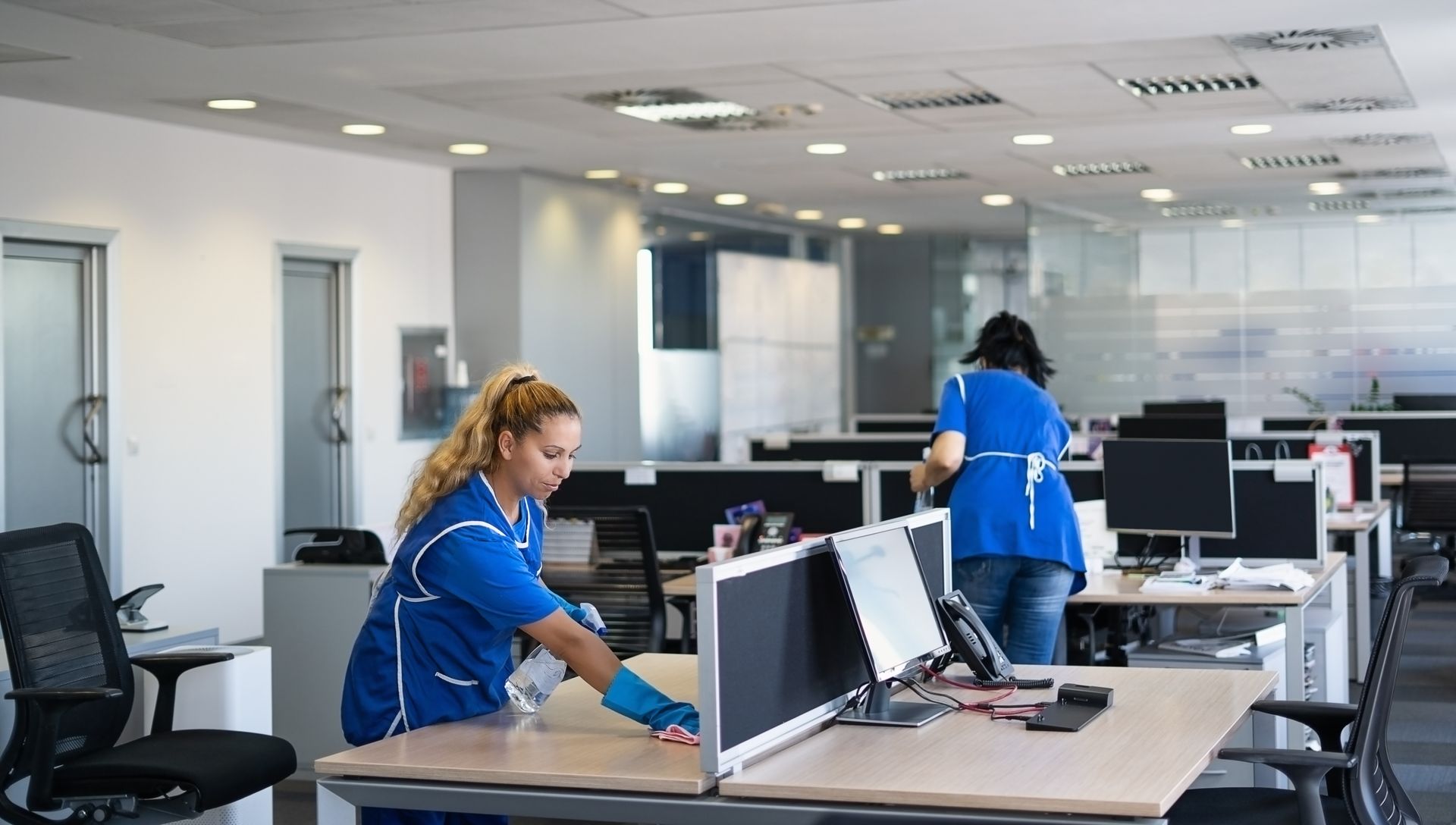 Two people cleaning an office. One sprays a desk, the other tidies up. Modern office with cubicles.