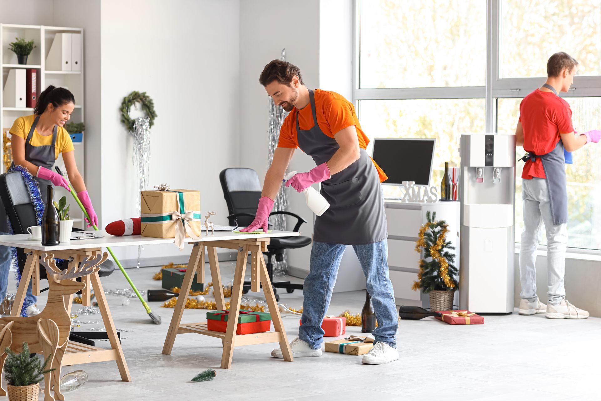 Three people cleaning an office. One sprays a desk, two wipe surfaces near a window.