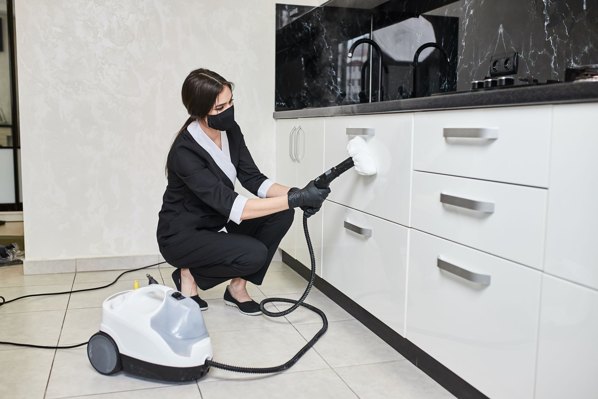 A woman is cleaning cabinets in a kitchen with a steam cleaner.
