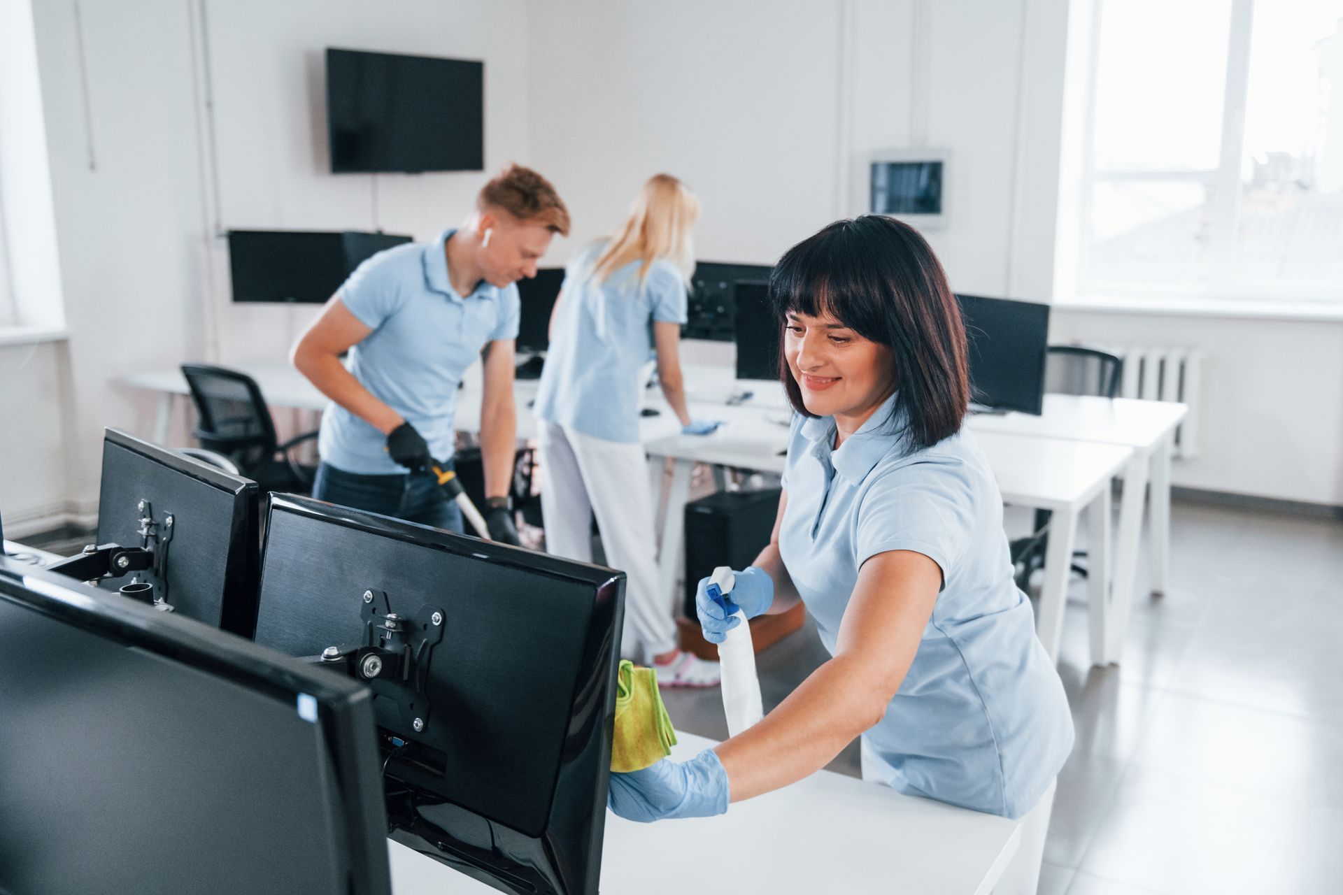 Cleaning crew sanitizing an office, wearing blue uniforms and gloves. A woman smiles while wiping a computer monitor.