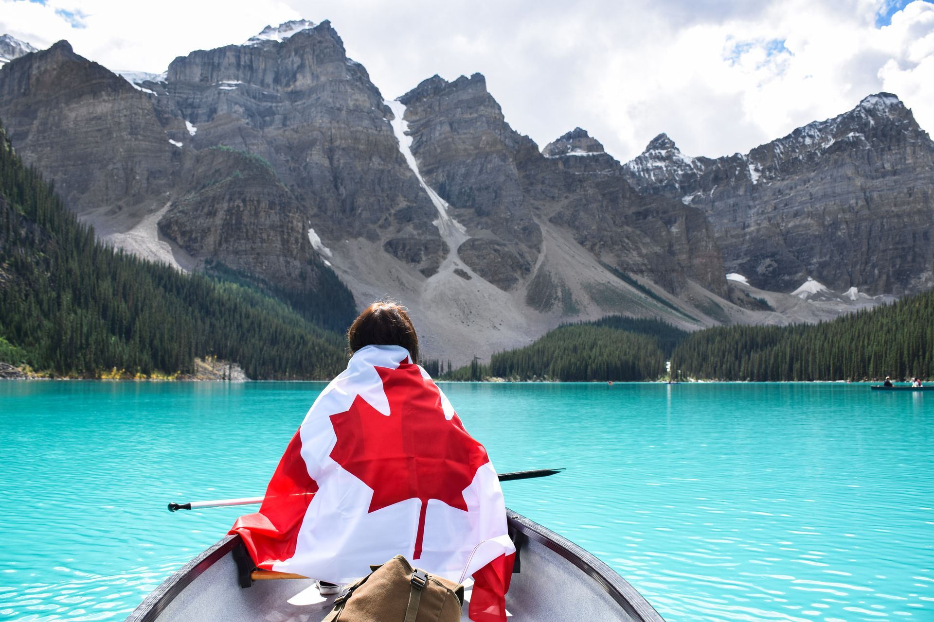 Person in canoe draped in Canadian flag, turquoise lake, mountain backdrop.