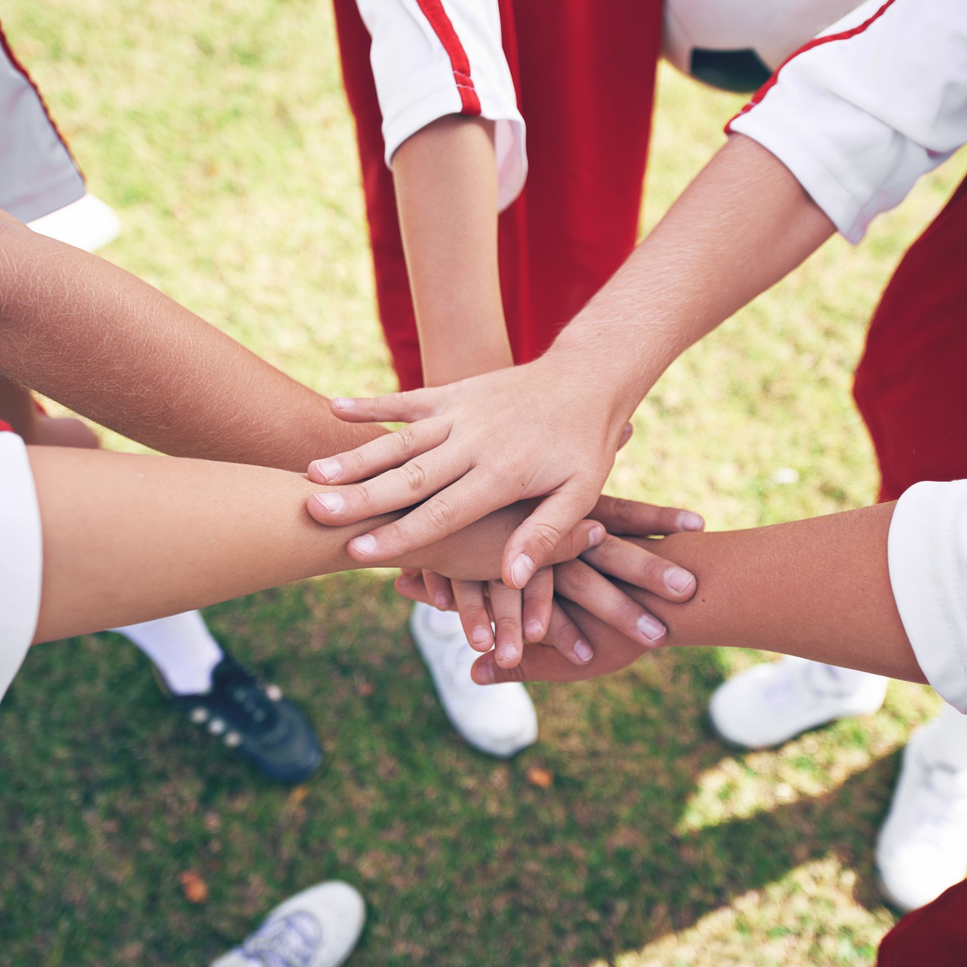 Hands of several people stacked in a team huddle, wearing red and white athletic uniforms, on grass.