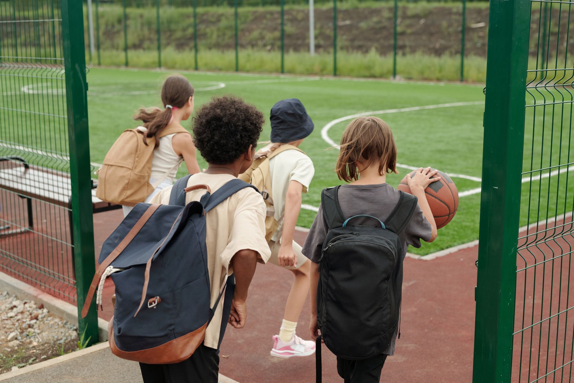 Children with backpacks walking toward a sports field, carrying a basketball. Green fence and grass in background.