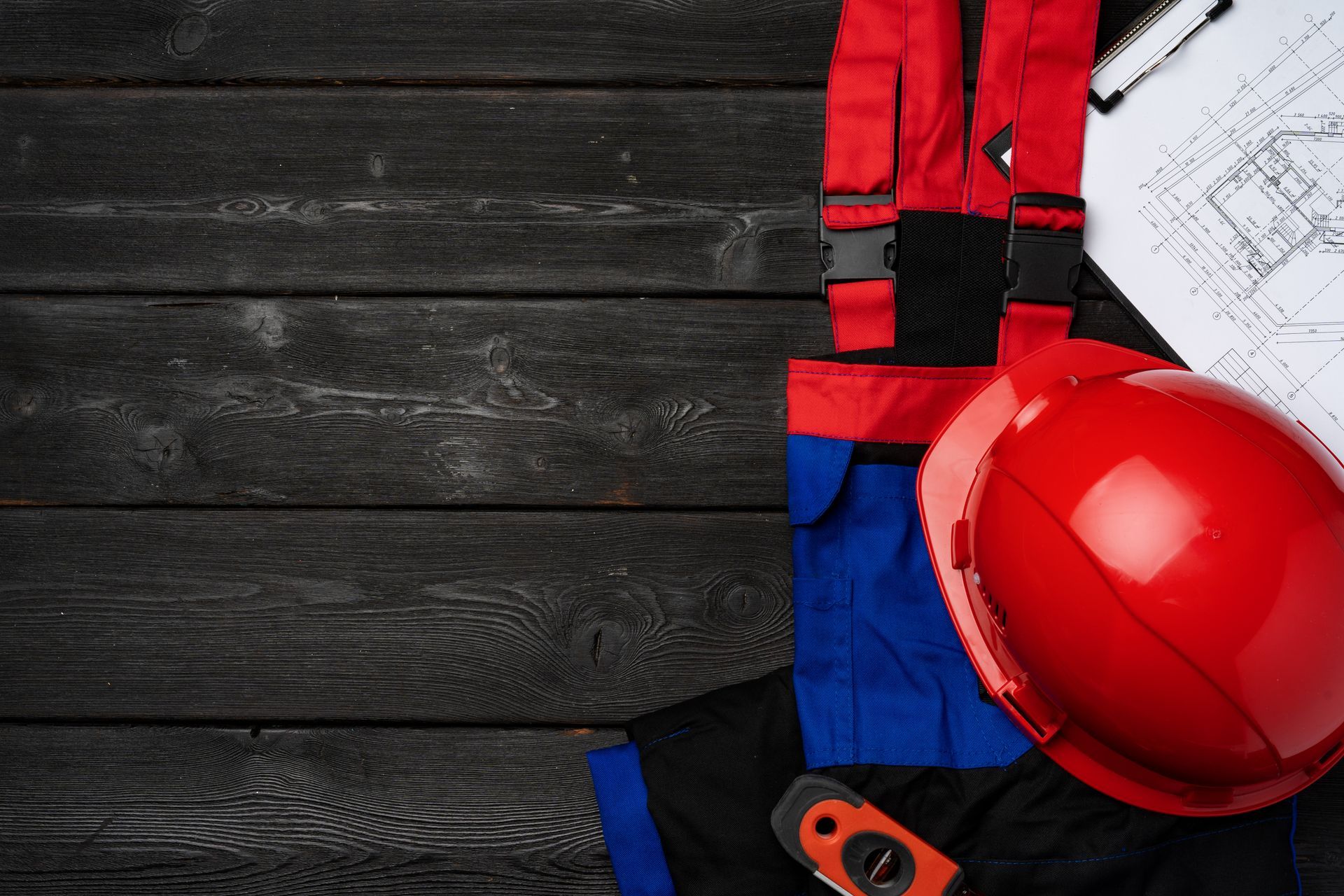 Red hard hat, work overalls, and blueprints on a dark wooden surface.