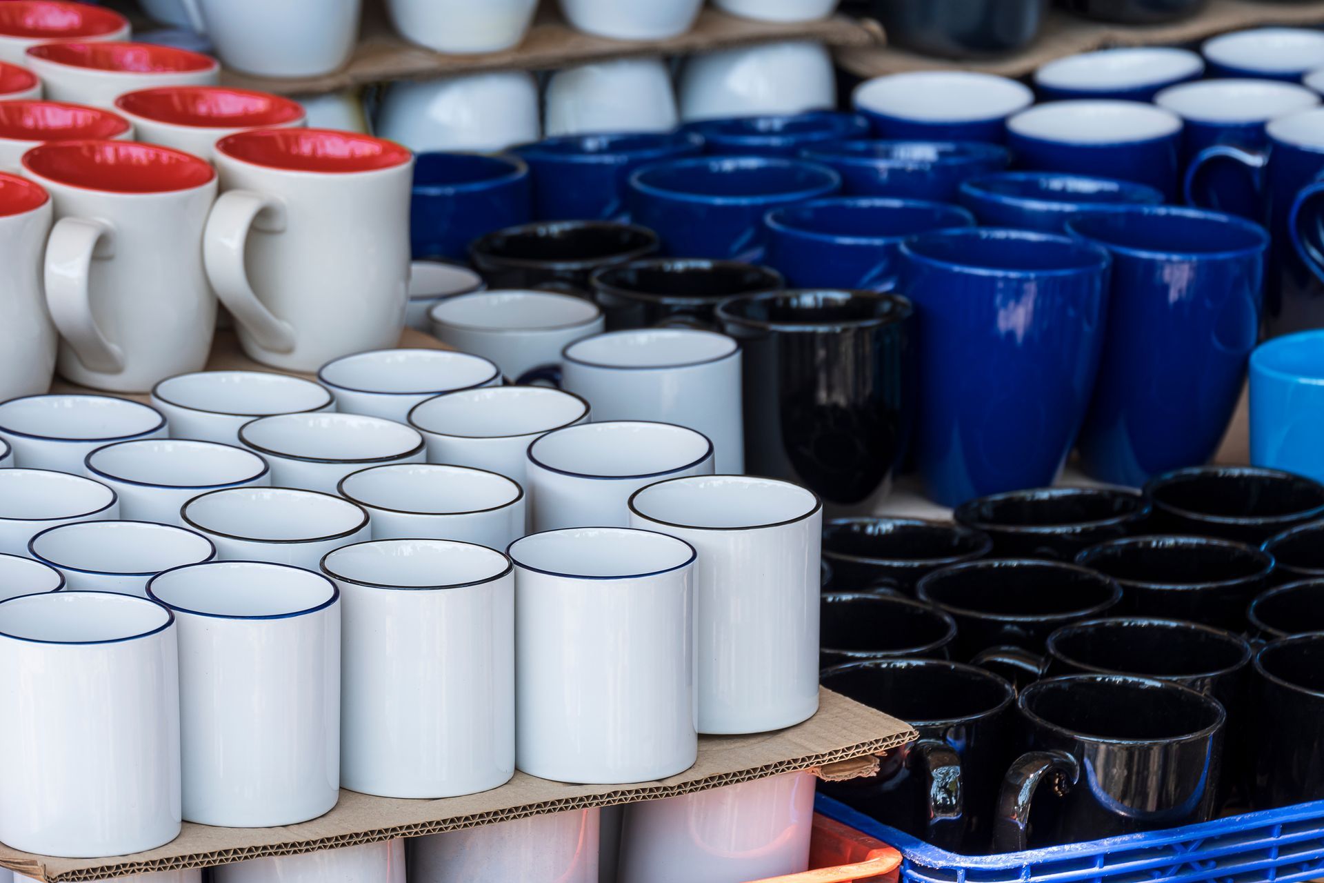 Rows of mugs in various colors, including white, red-lined, blue, and black, displayed for sale.