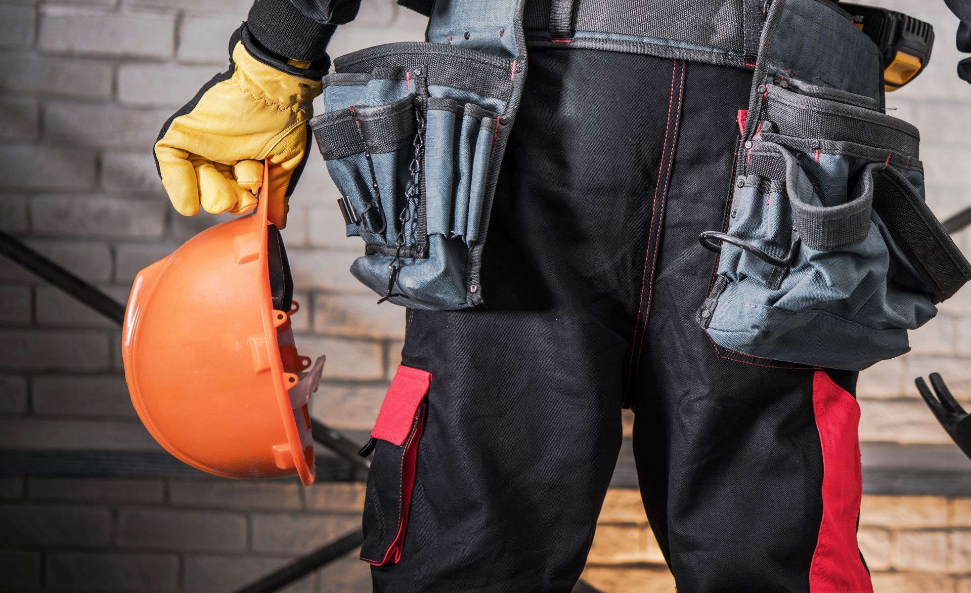 Construction worker holding orange hard hat, wearing tool belt and black work pants with red accents.
