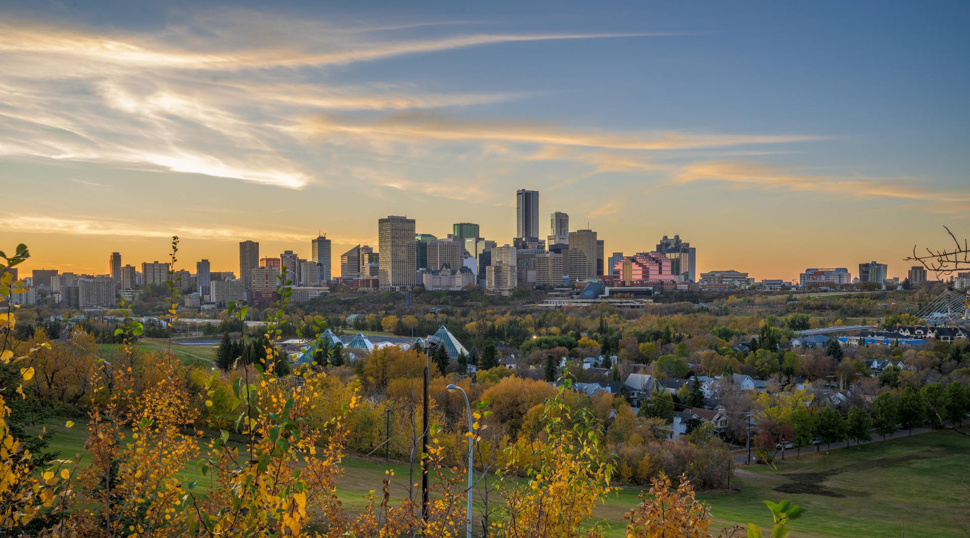 Skyline of a city at sunset with colorful clouds, trees, and buildings.