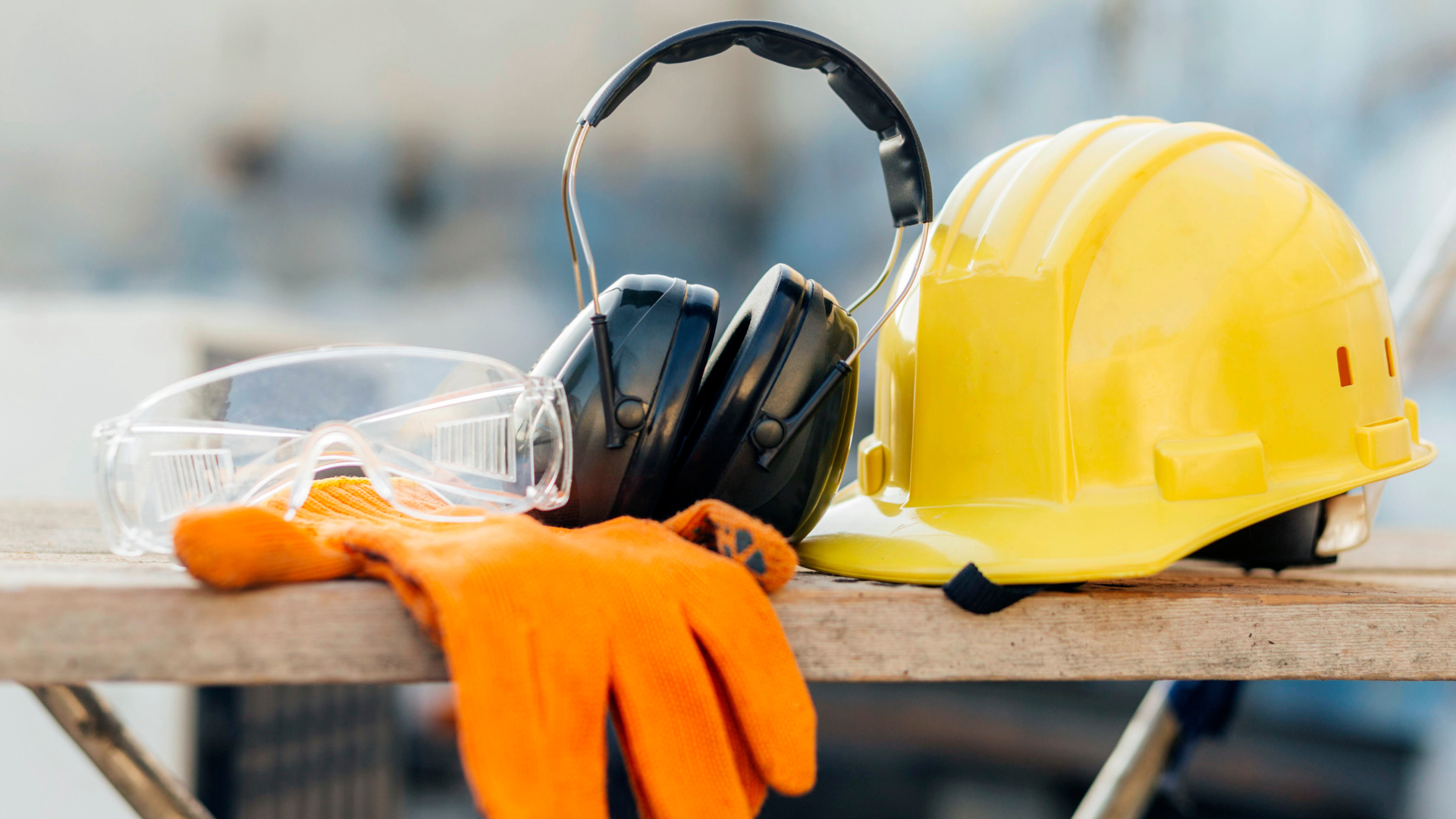Yellow hard hat, ear muffs, safety glasses, and orange work gloves on a wooden surface.