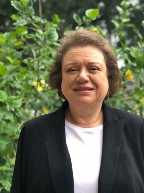 A woman in a black jacket and white shirt is standing in front of a tree.